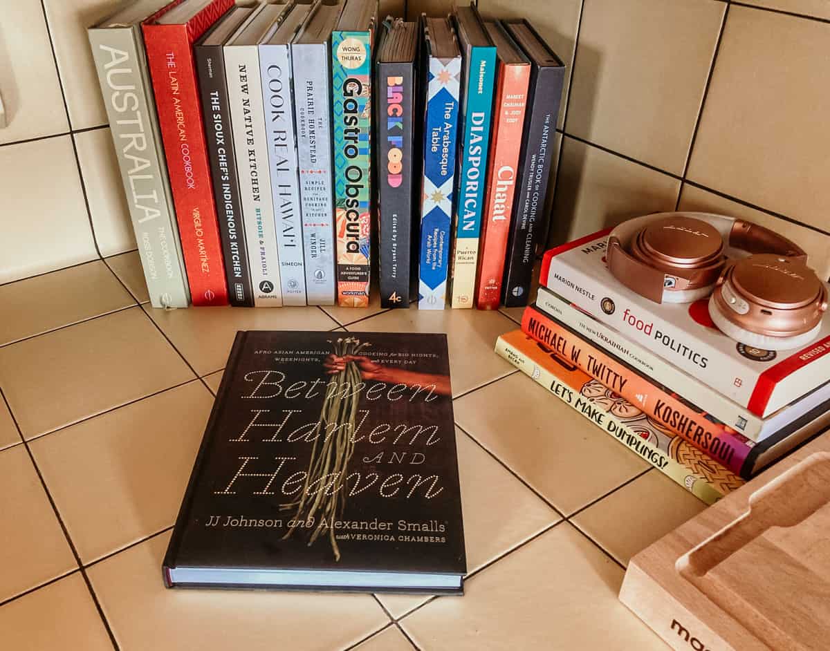 The cookbook Between Harlem and Heaven on a counter beside other cookbooks, food books, a pair of headphones, and a butcher block
